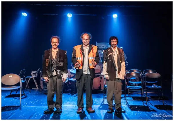 Escena de la obra 'Absurdia' con tres actores en un escenario minimalista lleno de sillas desordenadas, evocando un ambiente de misterio.