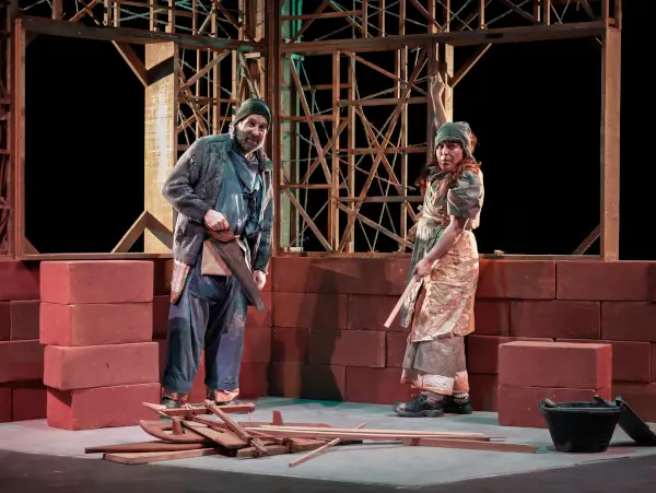 Actores en escena con elementos que sugieren una construcción, en una obra de teatro con tono surrealista y crítico.