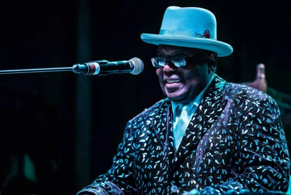 Kenny 'Blues Boss' Wayne tocando el piano con pasión en un escenario iluminado, con una expresión concentrada y enérgica.