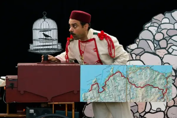 Un actor vestido con ropa tradicional gallega, sombrero y gafas, sentado en un triciclo antiguo con objetos y marionetas, en un escenario teatral.