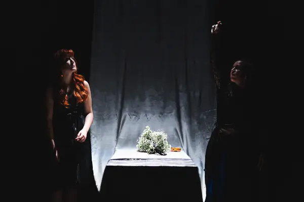 Escena de la obra 'Ninguén tira fotos nos enterros' con dos actrices en un escenario minimalista, una de ellas con una cámara antigua.