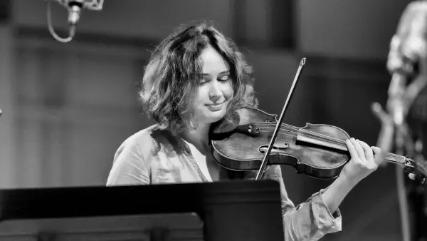 Patricia Kopatchinskaja, una violinista con expresión intensa, tocando su violín en el escenario con una orquesta de fondo.